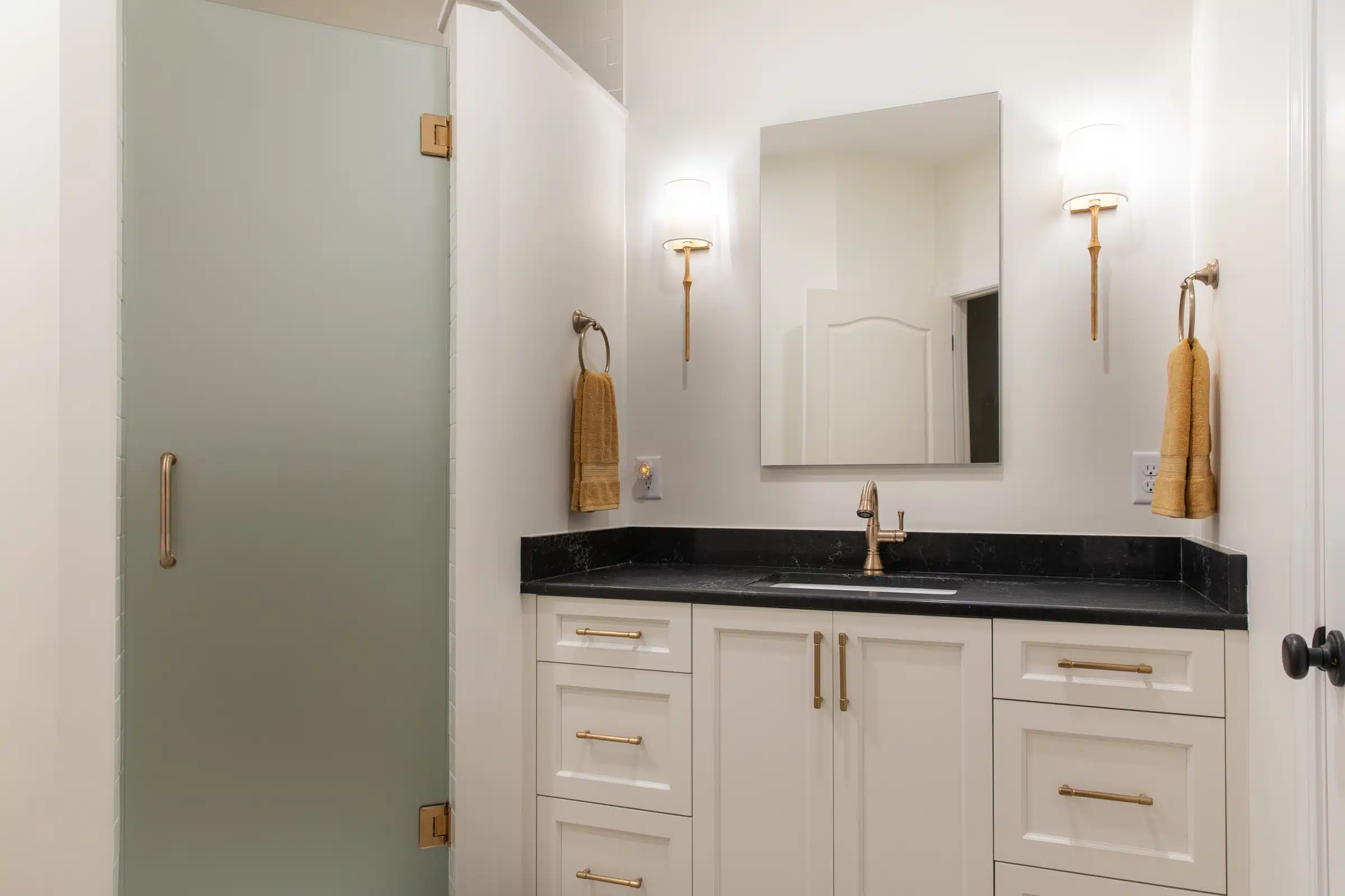 vanity with black stone top, brass fixtures, and frosted glass shower in DesRocher Hall